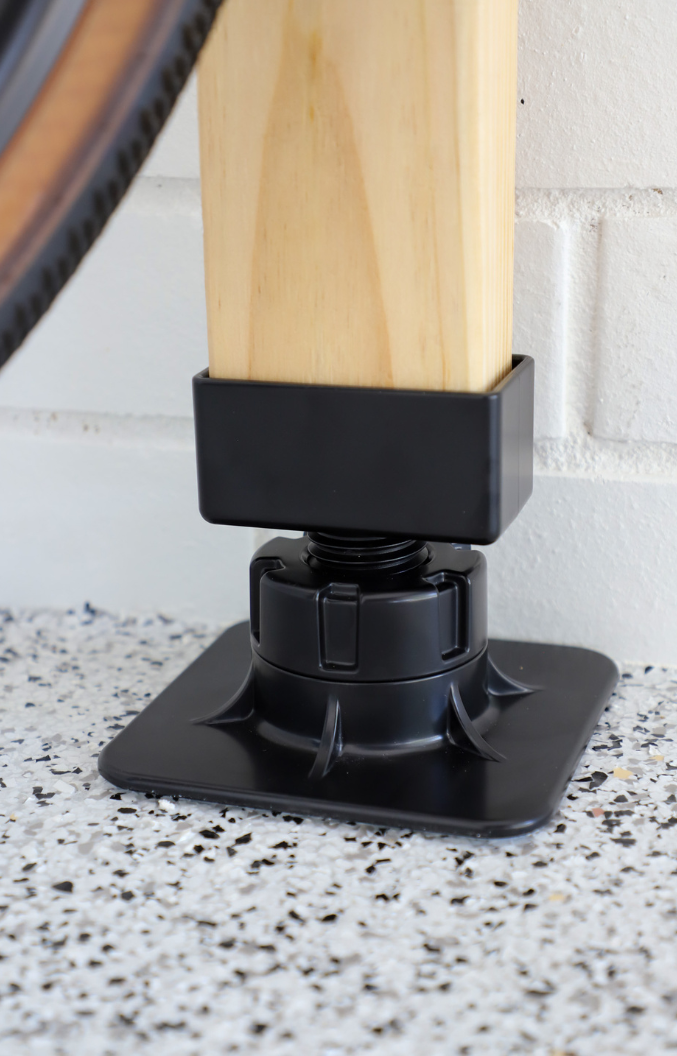 Black adjustable leg leveler on a wooden leg against a white brick wall.